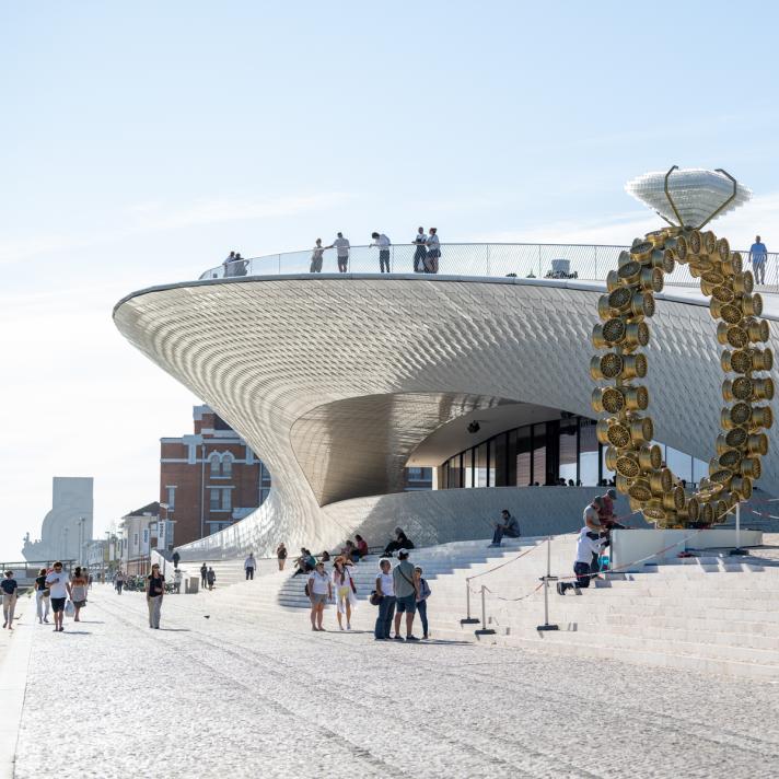 People walking in the sun at the riverside in front of the ew Museum of Art, Architecture and Technology (MAAT) in LIsbon - a wave shaped silver building.