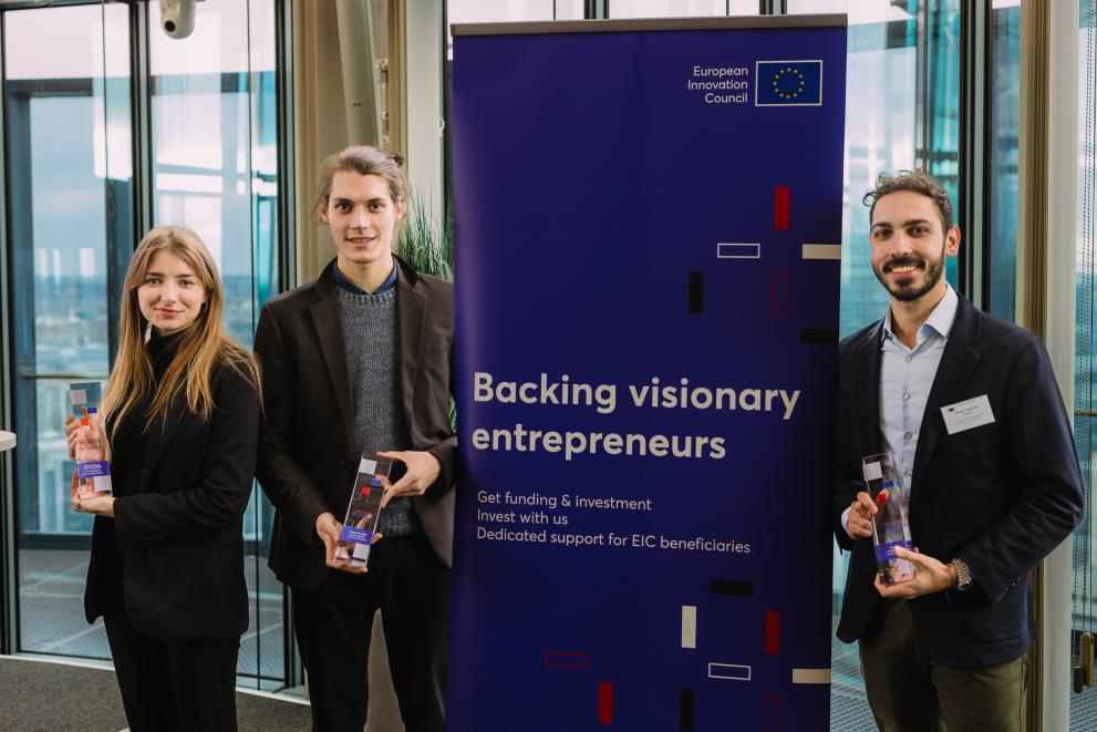 European Social Innovation Award winners Fiona Leiter, Ander Zabala Gomez, and Fabrizio Custorella proudly posing with their awards in front of the EIC roll-up that declares 'Backing up visionary entrepreneurs.